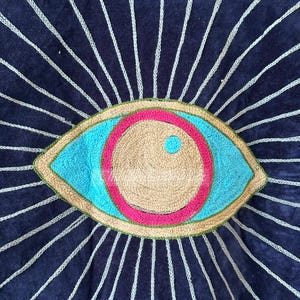 May include: A navy blue textile with an embroidered eye design. The eye features a light blue iris, a tan center, and a pink and green border. White stitched lines radiate from the eye, creating a sunburst effect.