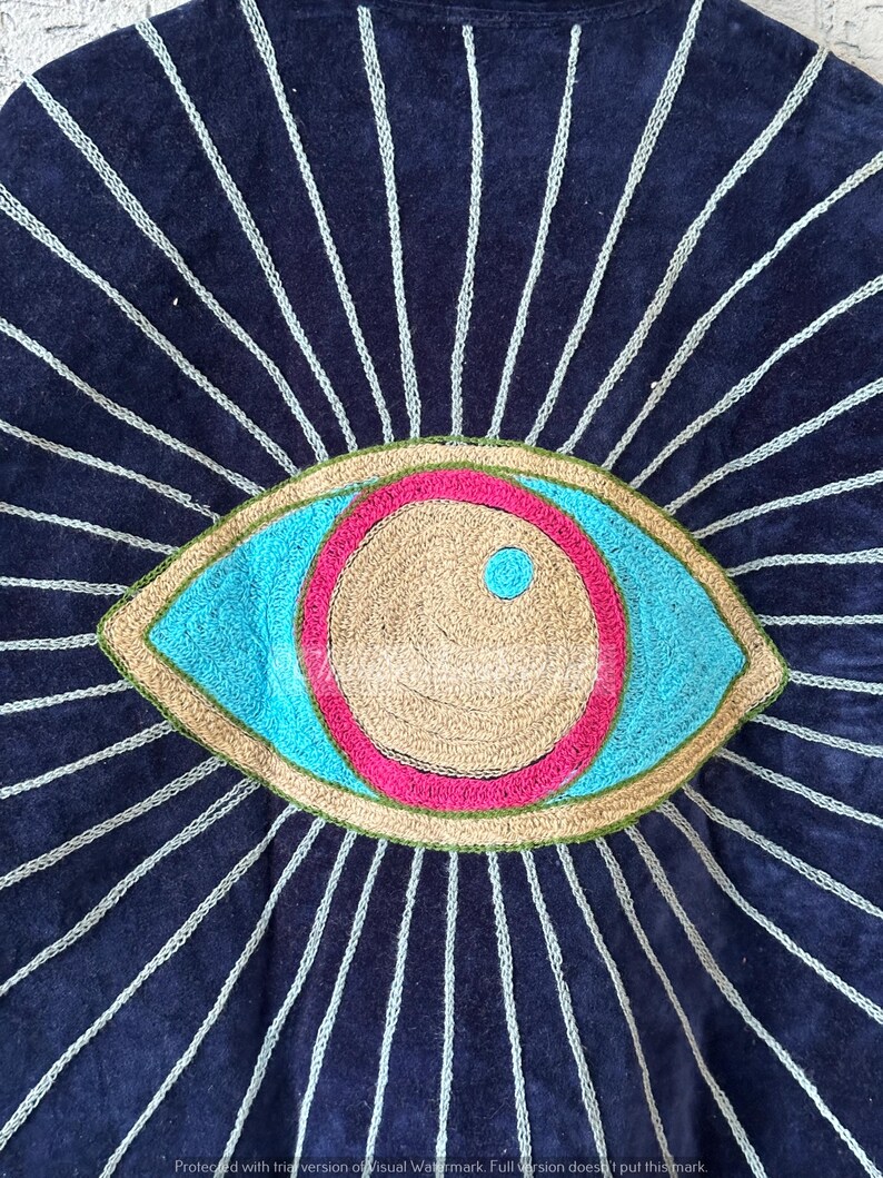 May include: A navy blue textile with an embroidered eye design. The eye features a light blue iris, a tan center, and a pink and green border. White stitched lines radiate from the eye, creating a sunburst effect.
