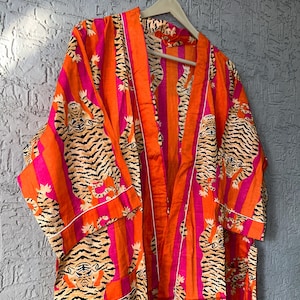 May include: An orange and pink striped robe with a tiger print. The robe has a white trim and is hanging on a wooden hanger. The robe is made of a lightweight fabric.