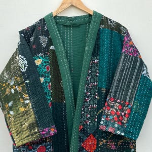May include: A patchwork kimono-style robe in shades of green, black, and floral patterns. The robe is made of various fabric patches sewn together with white stitching. It hangs on a wooden hanger against a white background.