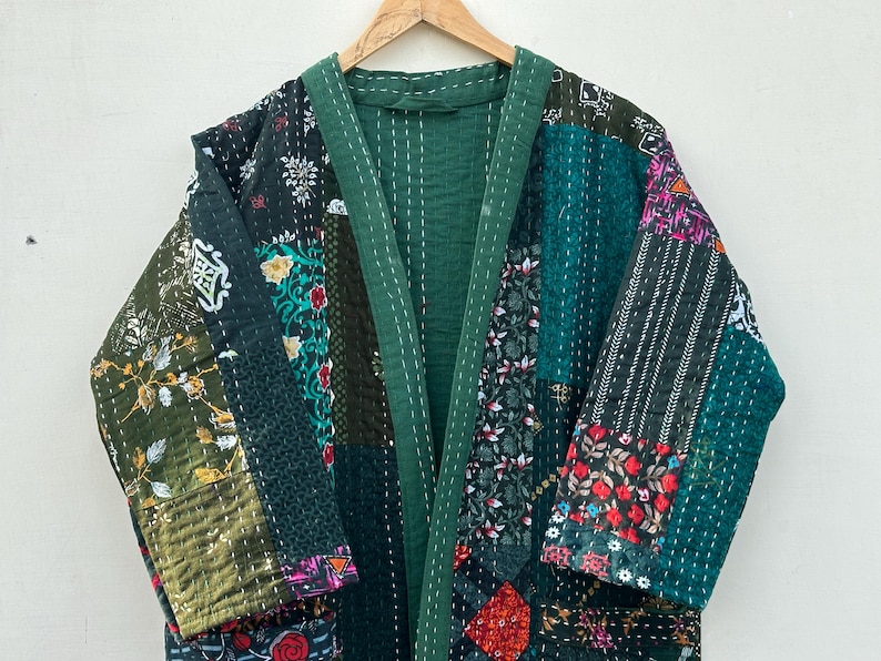 May include: A patchwork kimono-style robe in shades of green, black, and floral patterns. The robe is made of various fabric patches sewn together with white stitching. It hangs on a wooden hanger against a white background.