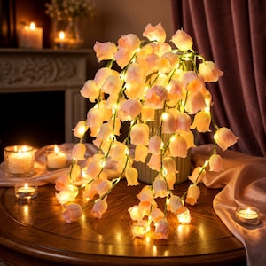 Puede incluir: Una lámpara decorativa con numerosas flores de color rosa pálido en forma de campana dispuestas en tallos verdes colgantes. Cada flor contiene una pequeña luz LED de tono cálido que crea un brillo suave. El arreglo floral se encuentra en un recipiente cilíndrico acanalado sobre una superficie de madera pulida.