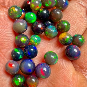 Op de afbeelding: Een verzameling gepolijste, ronde opalen in verschillende maten en kleuren. De opalen vertonen een kleurenspel, met flitsen van groen, blauw, rood en geel. De opalen worden in een hand gehouden, wat hun iriserende kwaliteiten laat zien.
