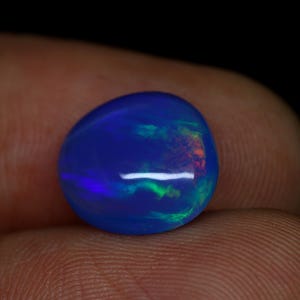 Peut inclure: Une pierre précieuse polie, de forme ovale, avec une couleur de base bleu foncé. La pierre présente des éclats de vert, d'orange et de violet, créant un jeu de couleurs. Le bijou est tenu entre les doigts sur fond sombre.