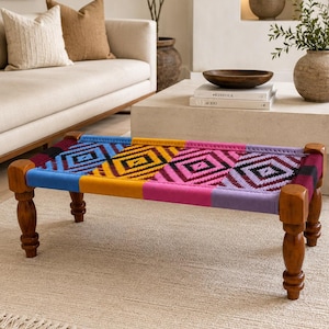 May include: A low rectangular bench with four turned wooden legs and a woven seat featuring a vibrant geometric pattern in blue, yellow, pink, and purple. The seat is divided into four distinct sections, each displaying a diamond motif.