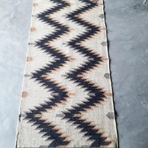 Può includere: Un tappeto rettangolare con sfondo color crema e un motivo a zig-zag ripetuto in blu navy, grigio e marrone. Il tappeto ha una frangia su entrambe le estremità. Questo tappeto decorativo è adatto per un corridoio o un ingresso.