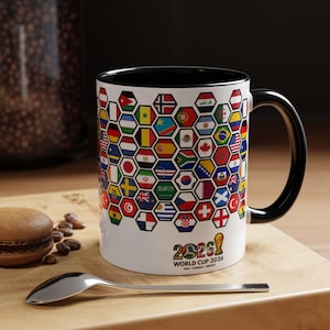 May include: White ceramic mug with a black interior and handle. The mug features a design of flags from around the world in a honeycomb pattern. The text "2026 WORLD CUP" is printed on the mug, along with the USA, Canada, and Mexico.