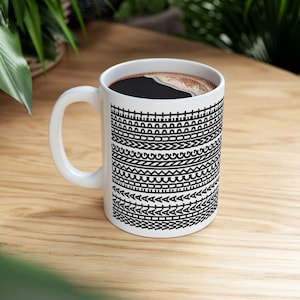 May include: A white ceramic mug filled with dark coffee, featuring a repeating black geometric pattern around the exterior. The design consists of horizontal bands of various shapes, including lines, semicircles, zigzags, and the repeated text WHAT.