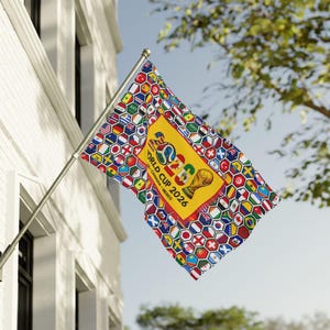 May include: A rectangular flag attached to a silver pole featuring a pattern of colorful hexagonal national flags surrounding a central yellow rectangle. The center displays the text WORLD CUP 2026 USA CANADA MEXICO and a golden trophy graphic.