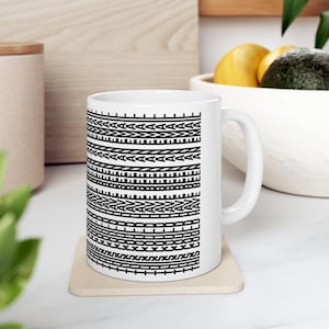 May include: A white ceramic mug featuring a repeating black geometric pattern that wraps around the exterior. The design consists of horizontal bands of triangles, chevrons, and linear motifs. The mug has a curved handle on the right side.