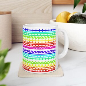 May include: A white ceramic mug featuring a repeating pattern of horizontal bands in vibrant rainbow colors. The design consists of various geometric shapes, including lines, zigzags, and arches, arranged in a stacked, colorful sequence.