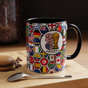 May include: A black-handled coffee mug featuring a design for the 2026 World Cup. The mug's exterior is covered in a pattern of hexagonal shapes, each containing a different national flag. The central design includes the World Cup trophy and the text "WORLD CUP 2026 USA - CANADA - MEXICO."