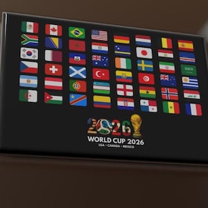 May include: A black rectangular plaque featuring the flags of various nations. The text "2026 WORLD CUP" is displayed below the flags, with "USA CANADA MEXICO" underneath. A golden trophy and soccer ball graphic are also present.