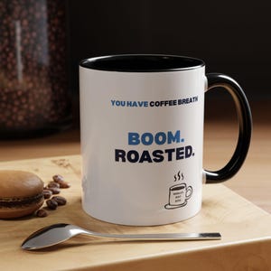 Puede incluir: Taza de café blanca con interior y asa negros. La taza tiene el texto "YOU HAVE COFFEE BREATH" y "BOOM. ROASTED." en azul. También se ve un pequeño gráfico de una taza de café con "WORLD'S BEST BOSS".
