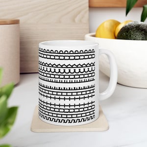 May include: White ceramic mug with a series of black geometric patterns. The patterns include arches, rectangles, and wavy lines. The mug has a standard handle and sits on a beige coaster. The mug is approximately 9.5 cm tall.
