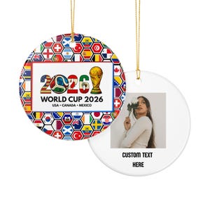 May include: Two round ceramic ornaments with gold hanging strings. One features a World Cup 2026 graphic with international flags in a honeycomb pattern, a soccer ball, and a trophy. The other displays a photo of a person holding a white flower.
