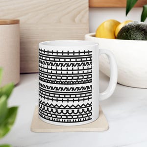 May include: A white ceramic mug featuring a repeating black geometric pattern consisting of horizontal bands of lines, dashes, squares, and semi-circles. The mug has a standard cylindrical shape with a curved handle on the right side and a smooth finish.