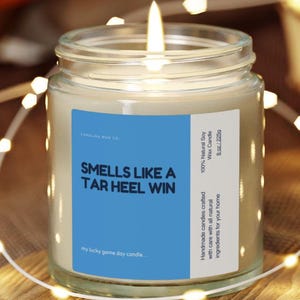 Könnte beinhalten: Eine brennende Sojawachskerze in einem Glas mit einem blauen Etikett mit der Aufschrift "Smells Like A Tar Heel Win". Die Kerze wiegt 225g und steht auf einer Holzoberfläche. Die Kerze ist von Lichterketten umgeben.
