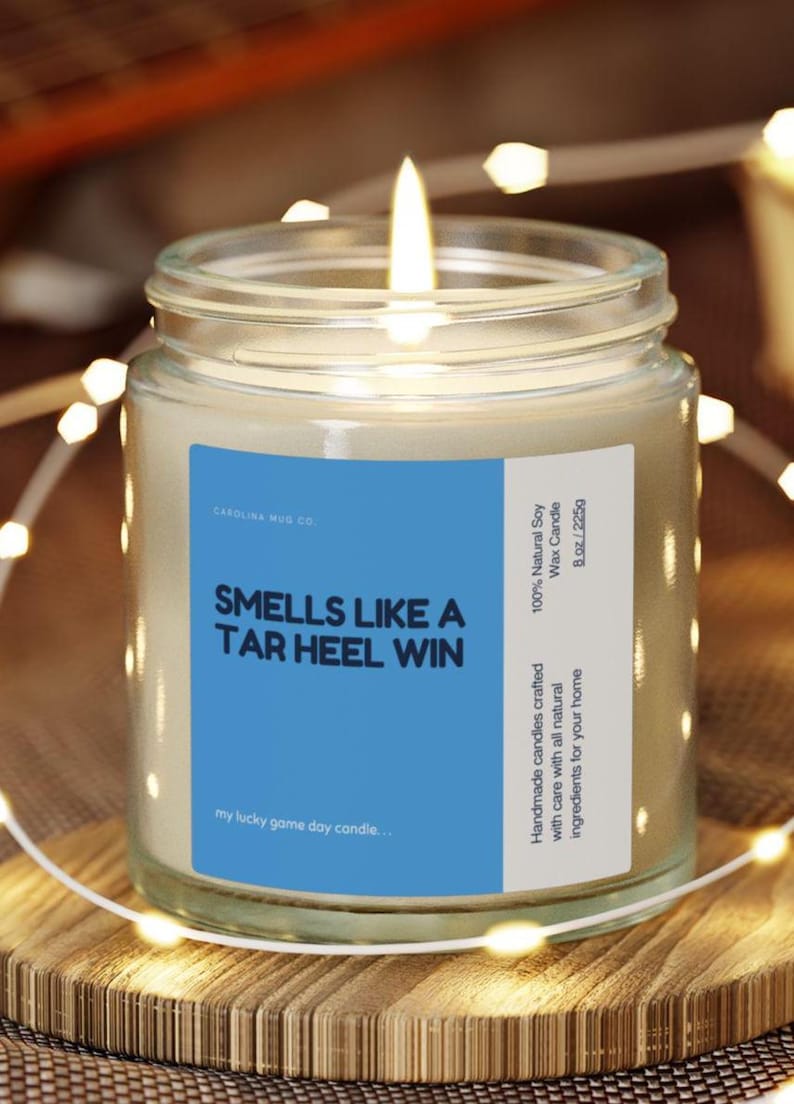Puede incluir: Una vela encendida de cera de soja en un tarro de cristal con una etiqueta azul que dice "Smells Like A Tar Heel Win". La vela es de 225g y est&aacute; sobre una superficie de madera. La vela est&aacute; rodeada de luces de cadena.
