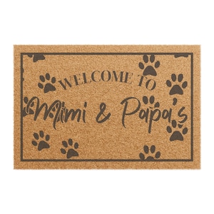 May include: A rectangular doormat made of natural coir fiber. The mat features a black border and the words "WELCOME TO Mimi & Papa's" in a script font, along with paw print designs. The background is a light brown color.