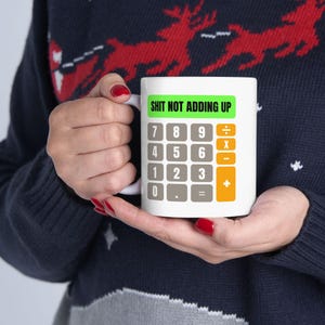 May include: White ceramic mug with a calculator design. The mug features a green rectangle with the text "SHIT NOT ADDING UP" above a calculator keypad. The mug is held by a person wearing a dark blue sweater.