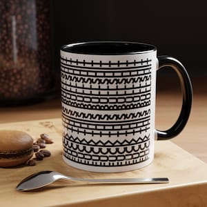 May include: A white ceramic mug with a black interior and handle, featuring a repeating horizontal pattern of black geometric lines, zigzags, and small arches encircling the exterior surface of the mug in a stark, high-contrast design.