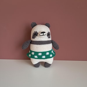 Könnte beinhalten: Handgemachte Panda-Bär-Puppe aus Häkelarbeit. Der Panda hat einen weißen Kopf und Körper mit schwarzen Akzenten an Ohren, Augen und Armen. Er trägt einen grünen Rock mit weißen Blumenmustern. Die Puppe ist etwa 15 cm groß.