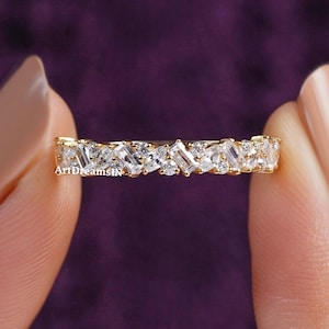 May include: A gold band ring featuring a continuous row of alternating baguette and round-cut clear gemstones. The ring is held between two fingers against a purple background. The text "ArtDreamsIN" is visible on the ring.
