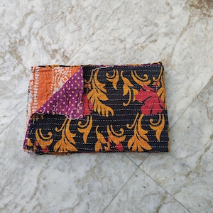 Könnte beinhalten: Eine rechteckige Kantha-Decke mit schwarzem Hintergrund, einem sich wiederholenden gelben Blumen- und Rankenmuster sowie roten Blumenakzenten. Eine Ecke ist umgeschlagen und zeigt ein violettes Innenfutter mit weißen Punkten und einem orangefarbenen Rand.