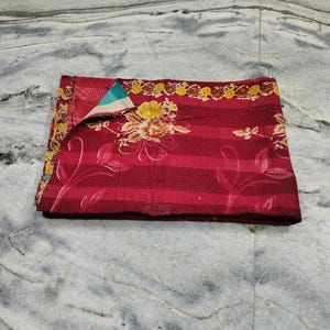 Könnte beinhalten: Ein gefaltetes rechteckiges Textil mit einer tiefroten Basis, horizontalen Streifen und einem auffälligen gelb-weißen Blumenmuster. Der Stoff zeigt durchgehend sichtbare Steppstichdetails, wobei eine Ecke umgeschlagen ist und ein blaugrünes Futter enthüllt.