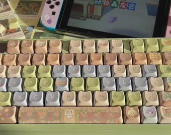 Puede incluir: Un teclado de colores pastel con teclas personalizadas con varios personajes y diseños de dibujos animados. Una Nintendo Switch rosa con un juego que se muestra en la pantalla se encuentra detrás del teclado. Las teclas son en tonos beige, verde y azul.