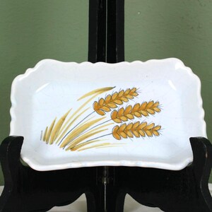Op de afbeelding: Een witte rechthoekige schaal met een geschulpte rand, met een tarwe-aar ontwerp in goud en bruin. De schaal wordt gepresenteerd op een zwart houten ezel tegen een groene achtergrond.
