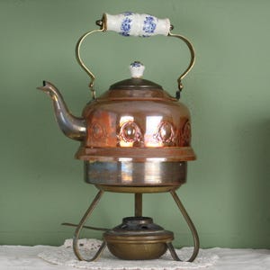 Op de afbeelding: Een koperen en messing theepot met een blauw-wit handvat, op een metalen standaard met een brander. De theepot staat op een tafel met boeken en een schilderij op de achtergrond.