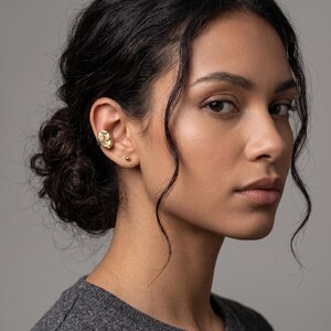 May include: A textured gold ear cuff and a small gold stud earring are visible on the ear. The person in the image has dark hair styled up and is wearing a grey sweater. The background is plain.