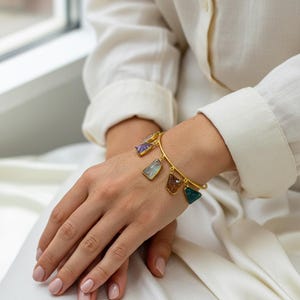 May include: Gold-toned bracelet featuring multiple gemstone charms in various shapes and colors. The bracelet is worn on a wrist, with the gemstones hanging freely. The gemstones include purple, blue, brown, and green hues.