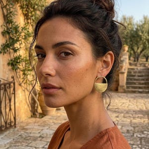 Puede incluir: Una persona lleva pendientes dorados en forma de semicírculo. Los pendientes tienen una superficie texturizada y un diseño sencillo y moderno. La persona lleva una blusa color óxido y tiene el pelo oscuro recogido. El fondo presenta un edificio de piedra y olivos.