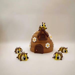 Op de afbeelding: Een bruine, bijenkorfvormige decoratie met een kleine deur, een dak en witte madeliefjesaccenten. Vier geel-zwarte bijenfiguren zijn rond de korf verspreid, met een bij bovenop. De achtergrond is wit.