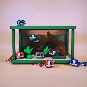 Peut inclure: Un diorama encadré de vert représentant une scène de forêt miniature avec une bûche marron et du feuillage vert. Plusieurs figurines de lézards colorés aux grands yeux sont disposées autour du diorama.
