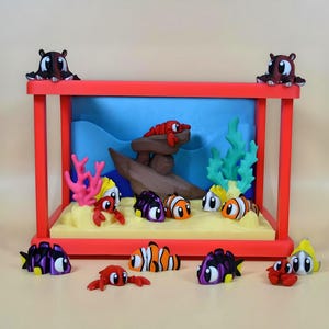 Puede incluir: Un diorama caprichoso con un marco rojo y una escena submarina en miniatura. La escena incluye un naufragio, corales y varios peces y cangrejos de dibujos animados en colores vibrantes. Dos figuras de pulpo se asientan sobre el marco.