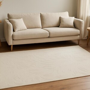 May include: A beige rug with a swirling pattern is placed on a hardwood floor. A beige sofa with two cushions and two throw pillows sits against a light-colored wall. A small wooden side table and a vase with dried plants are also visible.