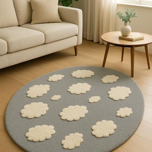 Pode incluir: Tapete cinza oval com formas de nuvem creme. O tapete está colocado sobre um chão de madeira, com um sofá bege e uma pequena mesa de madeira ao fundo. O design do tapete adiciona um toque lúdico.