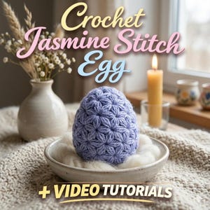 Op de afbeelding: Een lavendelgehaakt ei met een gestructureerd bloemmotief ligt in een kleine, crèmekleurige keramische kom gevuld met pluizig wit materiaal. De afbeelding bevat de tekst "Crochet Jasmine Stitch Egg" en "+ VIDEO TUTORIALS". Een brandende kaars en een vaas met droogbloemen staan op de achtergrond.