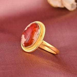 May include: A gold ring with an oval, red and white gemstone. The ring has a gold band and a bezel setting. The gemstone has a marbled appearance. The ring is set against a pink background. The ring's design is simple and elegant.