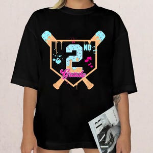 May include: Black t-shirt with a baseball-themed graphic. The design features a baseball diamond shape with crossed baseball bats, the number "2" in blue, and the word "Grade" in pink. The shirt is a casual style.