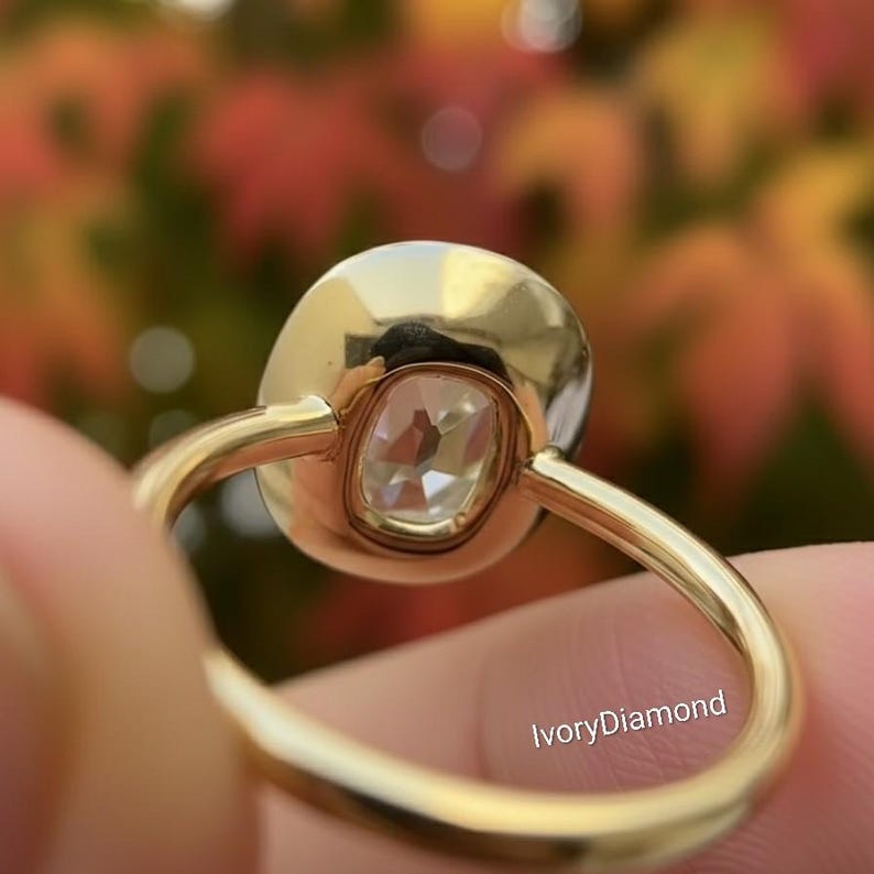 May include: Close-up of a gold ring with a large, clear, cushion-cut diamond. The ring has a simple, elegant design with a smooth, polished band. The text "IvoryDiamond" is visible on the ring.