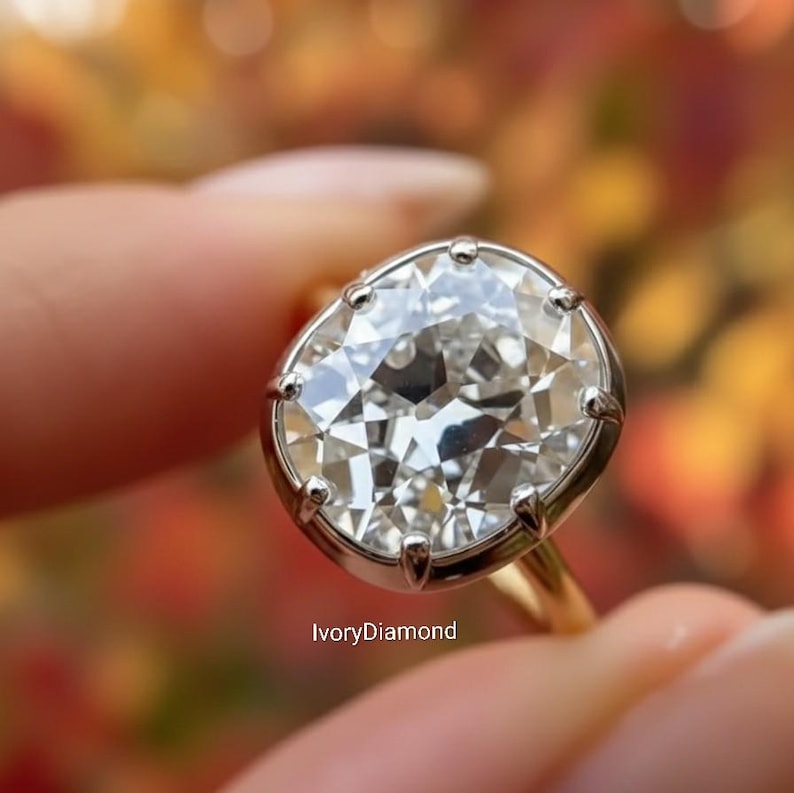 May include: A close-up of a diamond ring with a large, oval-shaped, clear diamond. The diamond is set in a silver bezel with a gold band. The ring is held by a person's fingers. The text "IvoryDiamond" is visible.