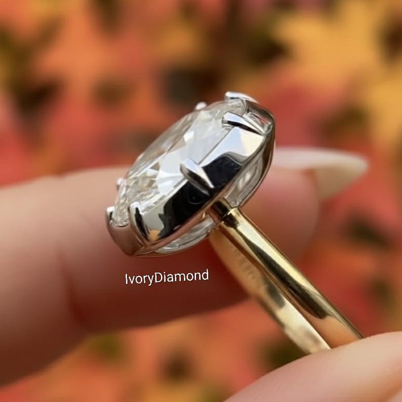 May include: Close-up of a diamond ring with a large, oval-cut gemstone. The ring has a two-tone design, with a gold band and a silver-colored setting around the diamond. The text "IvoryDiamond" is visible.