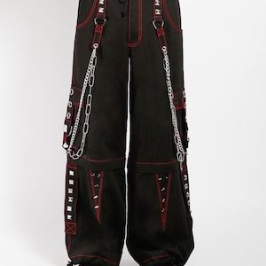 May include: Black cargo pants with red stitching and accents. The pants feature silver studs, chains, and straps. The design includes multiple pockets and a drawstring waist. The pants are wide-legged and have a punk or alternative aesthetic.