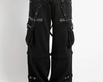 May include: Black cargo pants with multiple pockets, zippers, and metal grommet details. The pants have adjustable straps and a drawstring waist. The style is reminiscent of alternative fashion.