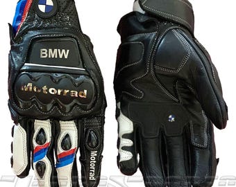 Może przedstawiać: Czarne skórzane rękawice motocyklowe z białymi i niebieskimi akcentami. Rękawice posiadają logo BMW i napis "Motorrad". Posiadają wzmocnione wyściółki i szwy dla ochrony.
